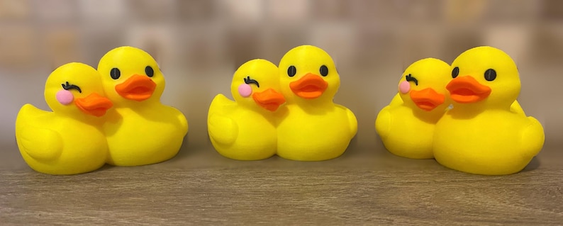 Three 3D printed couples Duck of a girlfriend and a boyfriend all yellows ducks connected with orange beaks and black eyes. Girlfriend duck has eyelashes and pink Rosie cheek. Rubber Duck style perfect for jeep ducking. Facing front.