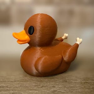 May include: A brown rubber duck shaped like a roasted turkey with a bright orange beak, black circular eyes, and two small white bone-shaped protrusions extending from the back, mimicking the appearance of drumsticks on a cooked poultry dish.