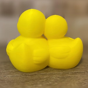 3D printed couples Duck of a girlfriend and a boyfriend all yellows ducks connected with orange beaks and black eyes. Girlfriend duck has eyelashes and pink Rosie cheek. Rubber Duck style perfect for jeep ducking. Facing back.