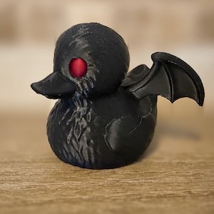 Puede incluir: Figura de pato negro con alas de murciélago y un ojo rojo. El pato tiene una superficie texturizada y un pico. Las alas están extendidas hacia los lados. La figura está sobre una superficie de madera.