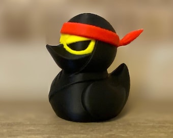 May include: A black rubber duck toy dressed as a ninja, with a red headband and yellow eye mask. The duck is on a wooden surface.