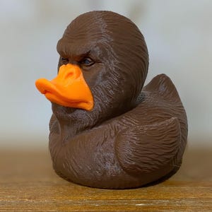 May include: A brown rubber duck with an orange beak and a stern, human-like face. The duck has detailed feather-like textures and is sitting on a wooden surface. The duck is a novelty item.