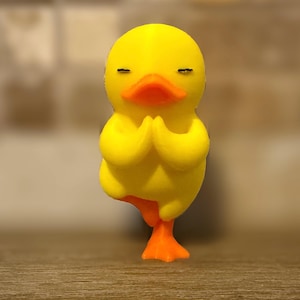 May include: A yellow duck figurine in a yoga pose. The duck has its eyes closed and its hands together in a prayer position. It is balancing on one orange foot. The duck's beak is orange.