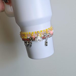 May include: A white tumbler adorned with multiple beaded bracelets. The bracelets feature yellow, pink, and clear beads, along with silver charms of elephants, a circus tent, and a peanut. The bracelets are stacked on the tumbler.