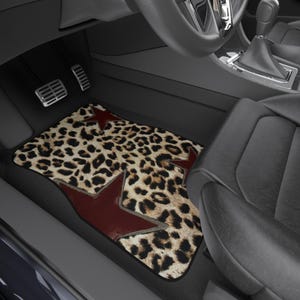 May include: A rectangular car floor mat featuring a tan and black leopard print pattern accented with three dark red stars of varying sizes. The mat is positioned on the floor of a vehicle interior, situated between the pedals and the seat.