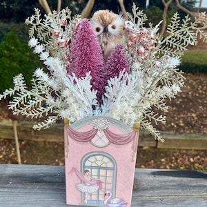 May include: A decorative pink container with a ballerina and swan illustration, filled with an owl figurine, pink bottle brush trees, and white and silver decorative branches. The container has a decorative arch top.