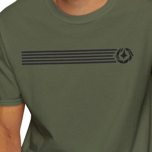 May include: Olive green t-shirt featuring a horizontal design of five parallel black lines and a circular emblem on the right side. The shirt has short sleeves and a crew neck.