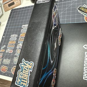 May include: Black card binder with stickers. The binder has the text "THE FIRST CHAPTER" on the top and "AZURITE SEA" on the side. Other stickers with text and graphics are also visible. The binder is on a gray cutting mat.