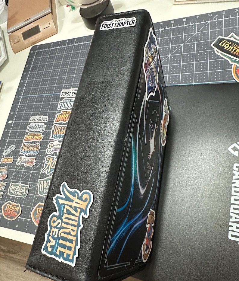 May include: Black card binder with stickers. The binder has the text "THE FIRST CHAPTER" on the top and "AZURITE SEA" on the side. Other stickers with text and graphics are also visible. The binder is on a gray cutting mat.