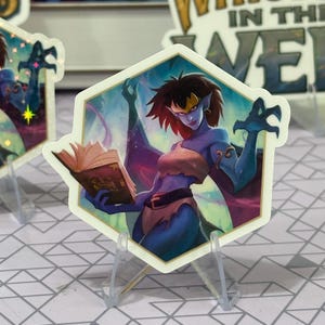May include: A hexagonal sticker featuring a blue-skinned character with red hair, holding a book. The character is wearing a tan top and a brown belt. The sticker has a gold border and is displayed on a clear stand.