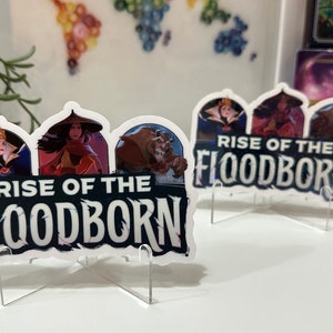 K&ouml;nnte beinhalten: Zwei wei&szlig;e Acryl-St&auml;nder mit einem gedruckten Bild von drei Disney-Charakteren, der b&ouml;sen K&ouml;nigin, Mulan und dem Biest, mit dem Text "Rise of the Floodborn".