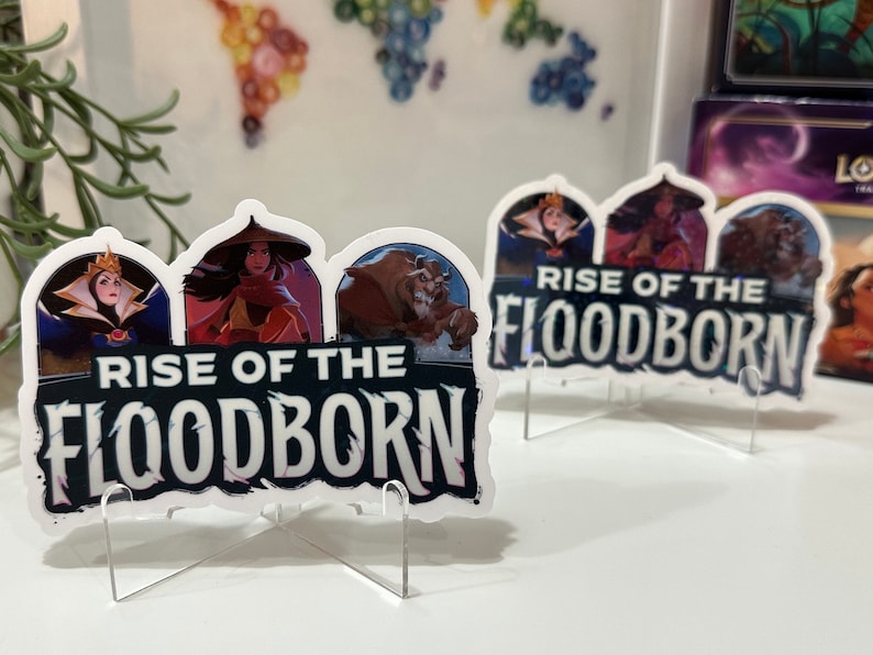 K&ouml;nnte beinhalten: Zwei wei&szlig;e Acryl-St&auml;nder mit einem gedruckten Bild von drei Disney-Charakteren, der b&ouml;sen K&ouml;nigin, Mulan und dem Biest, mit dem Text "Rise of the Floodborn".