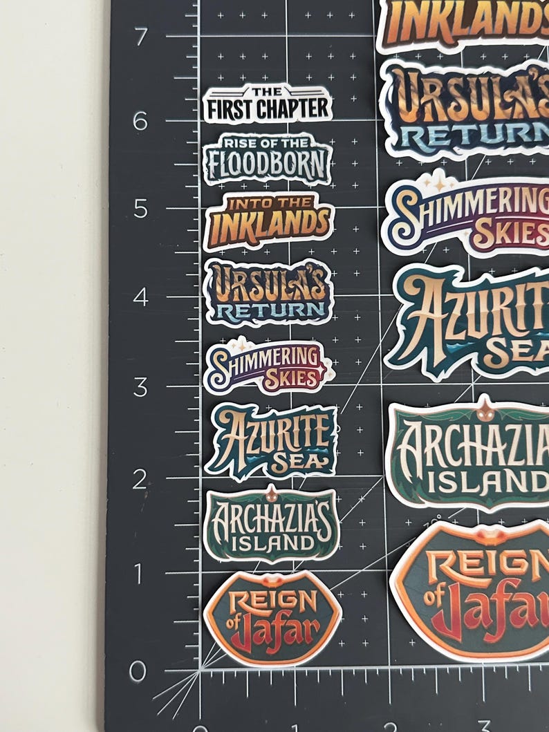 May include: A collection of colorful stickers with various fantasy-themed text labels. The stickers feature phrases like "The First Chapter," "Rise of the Floodborn," and "Ursula's Return." The stickers are arranged on a black surface with measurement markings.