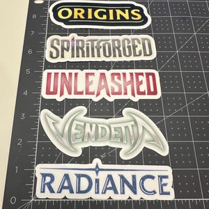 May include: Five rectangular stickers with rounded edges featuring the words ORIGINS in yellow, SPIRITFORGED in grey, UNLEASHED in red, VENDETTA in silver with stylized lettering, and RADIANCE in blue, all arranged on a black cutting mat with inch markings.