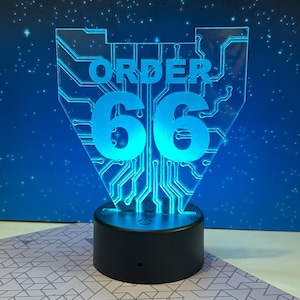 May include: A blue LED lamp with the words "ORDER 66" in a circuit board design. The lamp has a black base and is illuminated against a blue background with a starry pattern.