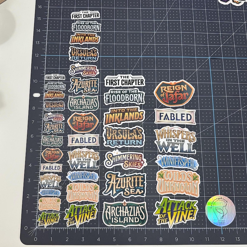 May include: A collection of colorful, die-cut stickers with various fantasy-themed text and designs. The stickers feature phrases like "First Chapter," "Floodborn," and "Ursula's Return." The stickers are arranged on a black grid.