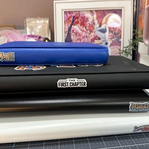 May include: A stack of four binders, with a blue binder on top, followed by black and white binders. The blue binder has the text "Azurite Sea". The black binder has the text "The First Chapter" and "Ursula's Return". The white binder has the text "Shimmering Skies".