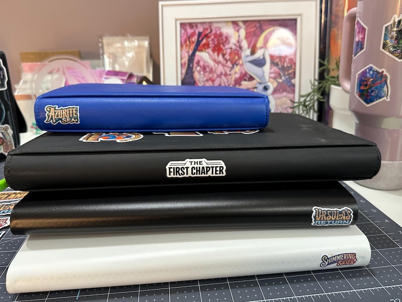 May include: A stack of four binders, with a blue binder on top, followed by black and white binders. The blue binder has the text "Azurite Sea". The black binder has the text "The First Chapter" and "Ursula's Return". The white binder has the text "Shimmering Skies".