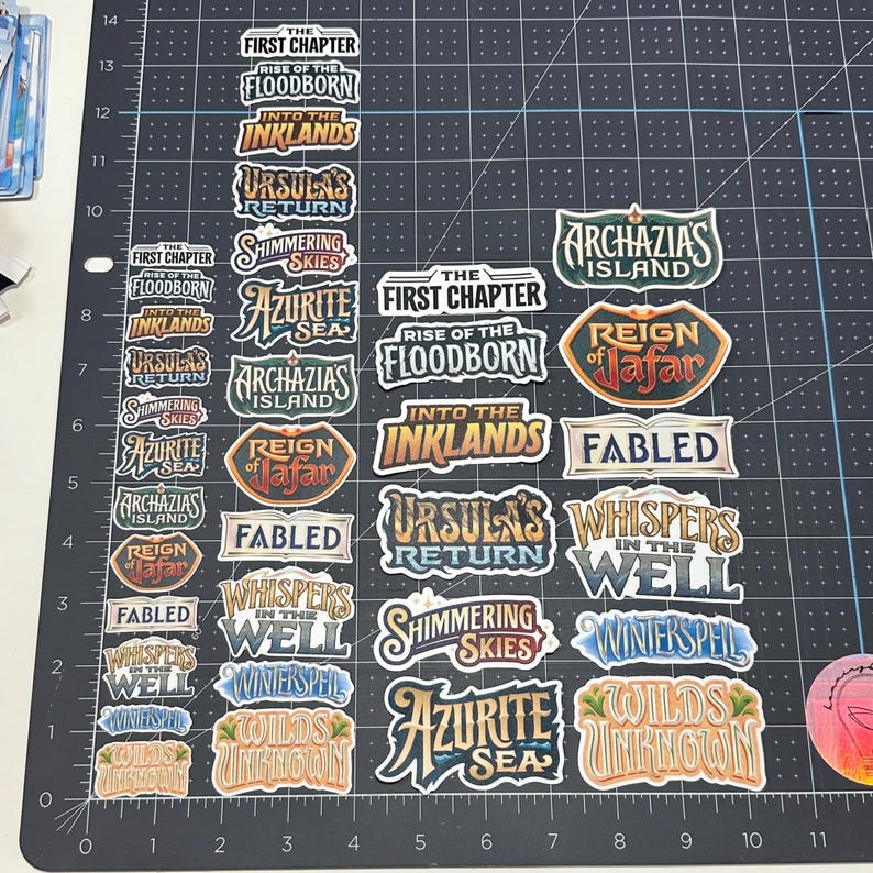 May include: A collection of colorful stickers with various titles, including "The First Chapter," "Rise of the Floodborn," and "Into the Inklands." The stickers feature ornate fonts and designs, with a mix of blues, greens, and browns. The stickers are arranged on a black grid.