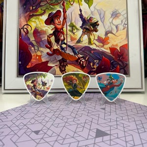 Può includere: Un'opera d'arte incorniciata con personaggi del gioco di carte collezionabili Lorcana di Disney, con tre plettri in primo piano. I plettri mostrano illustrazioni colorate di personaggi Disney. L'opera d'arte e i plettri sono su una superficie a motivi.