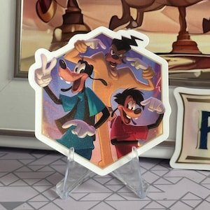 Puede incluir: Una pegatina hexagonal con tres personajes de dibujos animados en colores vibrantes. Un personaje lleva una chaqueta verde azulado, otro una camisa amarilla y el tercero una camisa roja. Los tres están señalando con los dedos. La pegatina se muestra en un soporte de plástico transparente.