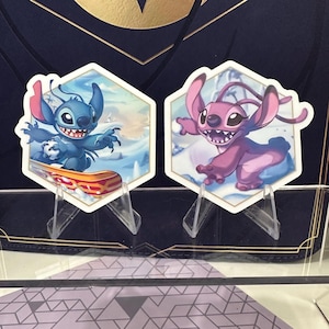 May include: Two hexagonal stickers featuring cartoon characters. One shows a blue creature on a snowboard, the other a pink creature. Both have white borders and are displayed on clear stands. The background depicts a snowy landscape.