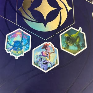 May include: Three hexagonal stickers featuring cartoon characters. One shows a blue alien in a bubble, another features Woody and Buzz Lightyear, and the third depicts a green Tyrannosaurus Rex. The stickers are set against a dark blue background.