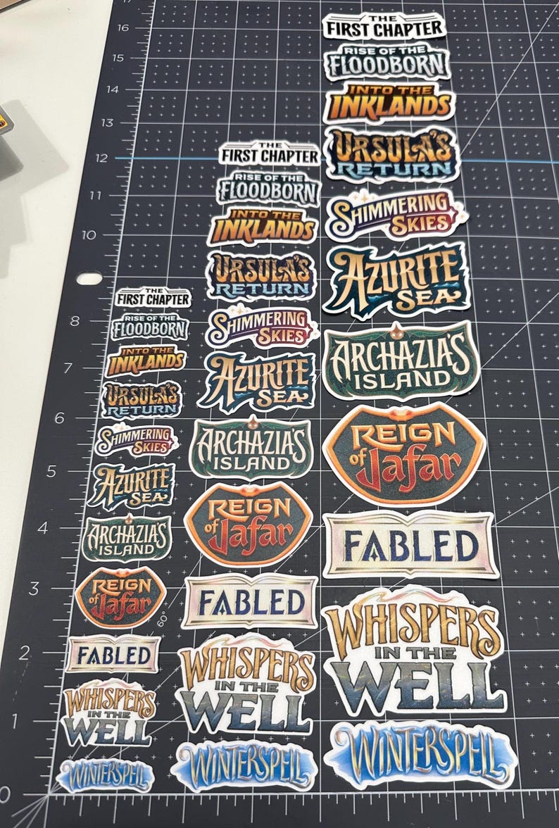 May include: A collection of colorful stickers with various fantasy-themed text labels. The stickers include phrases like "The First Chapter," "Rise of the Floodborn," and "Whispers in the Well." The stickers are arranged on a black cutting mat.
