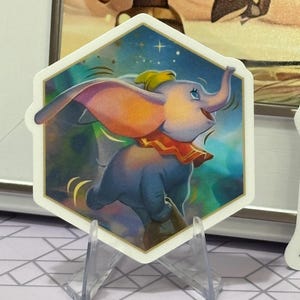 May include: A hexagonal sticker featuring Dumbo the elephant from the Disney film. Dumbo is depicted with large ears, a yellow hat, and a red collar, set against a blue and gold background. The sticker is displayed on a clear stand.