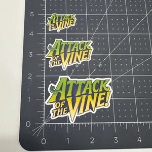 May include: Three stickers with the text "ATTACK OF THE VINE!" in a comic book style. The stickers are white with green, yellow, and blue lettering. The stickers are on a black cutting mat with a ruler.
