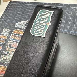 May include: A black binder with a sticker that reads "Archazias Island" in a green and white design. Several other stickers with text are visible, including "Floodborn" and "Into the Inklands". The binder is on a gray cutting mat.
