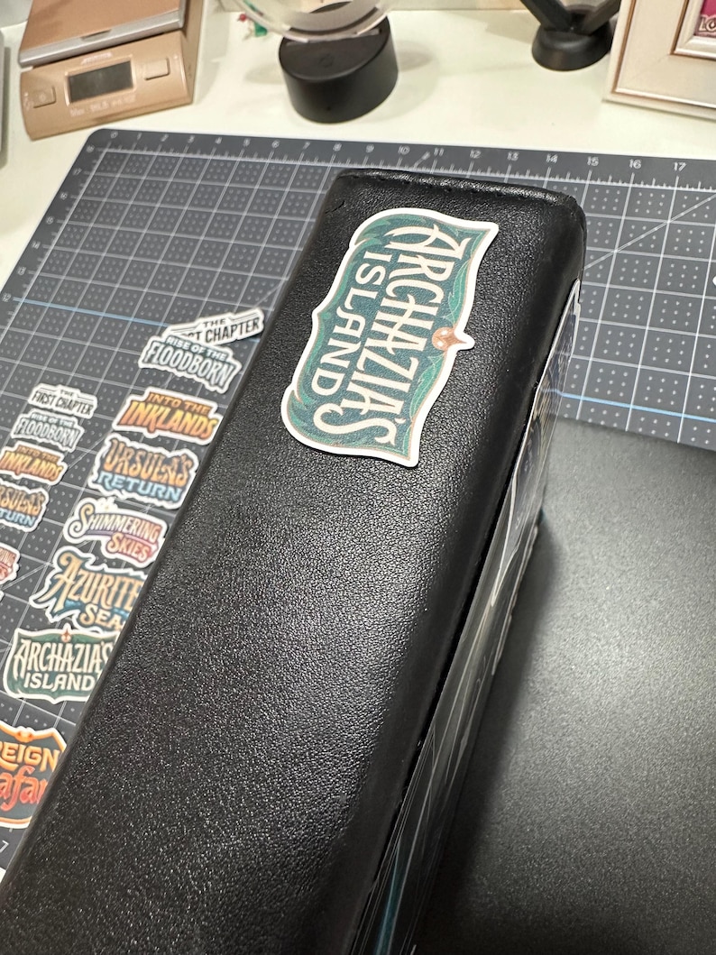 May include: A black binder with a sticker that reads "Archazias Island" in a green and white design. Several other stickers with text are visible, including "Floodborn" and "Into the Inklands". The binder is on a gray cutting mat.