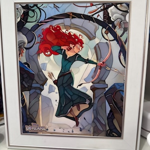 May include: A framed print featuring a character with long red hair wearing a dark green dress, holding a bow and arrow in a dynamic pose. The background shows stone ruins and foliage. The Disney Lorcana trading card game logo is in the corner.