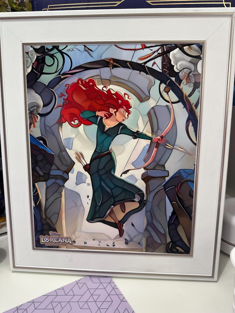 May include: A framed print featuring a character with long red hair wearing a dark green dress, holding a bow and arrow in a dynamic pose. The background shows stone ruins and foliage. The Disney Lorcana trading card game logo is in the corner.