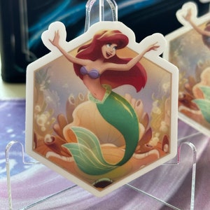 May include: A hexagonal sticker featuring Ariel, the Little Mermaid, in a seashell with a green tail and red hair. The sticker is on a clear acrylic stand.