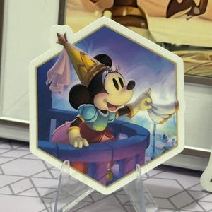 Könnte beinhalten: Ein sechseckiger Aufkleber mit Minnie Mouse in einem Prinzessinnen-Outfit. Minnie trägt einen goldenen Hut und ein blau-rosa Kleid und hält ein weißes Tuch. Der Hintergrund zeigt eine Schlossszene.