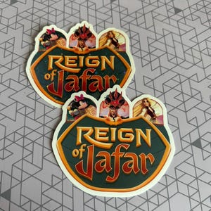 K&ouml;nnte beinhalten: Zwei Aufkleber mit gr&uuml;nem Hintergrund und goldener Schrift, die "Reign of Jafar" lautet. Die Aufkleber zeigen Comicbilder von Jafar, Jasmin und Aladdin.