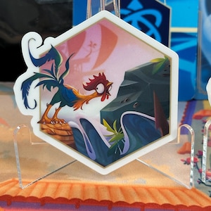 May include: A hexagonal sticker featuring a cartoon rooster with red wattle and comb, standing on a rock formation. The rooster is facing left and looking at a large, gray rock with green foliage. The background is a pink and orange gradient with a white outline.