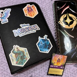 May include: A black binder with the Disney Lorcana Trading Card Game logo. The binder has several colorful stickers on it, including a blue bird, a blue and white character, a purple character, and a yellow character. The binder is open to reveal a card with a Winnie the Pooh character.