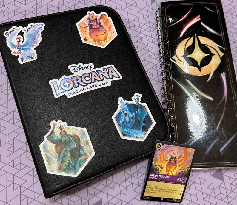 May include: A black binder with the Disney Lorcana Trading Card Game logo. The binder has several colorful stickers on it, including a blue bird, a blue and white character, a purple character, and a yellow character. The binder is open to reveal a card with a Winnie the Pooh character.