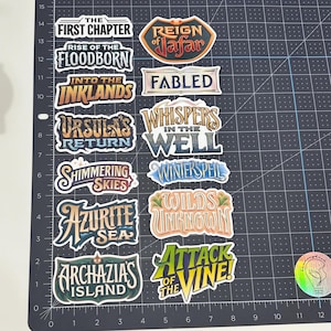 May include: A collection of colorful stickers with various fantasy-themed text designs. The stickers feature phrases like "The First Chapter," "Reign of Jafar," and "Ursula's Return." The stickers are arranged on a black cutting mat.