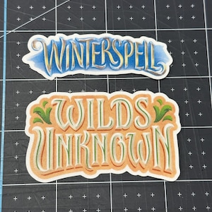 May include: Two decorative stickers with text. The top sticker reads "WINTERSPELL" in gold lettering with a blue and yellow background. The bottom sticker reads "WILDS UNKNOWN" in orange and green lettering.
