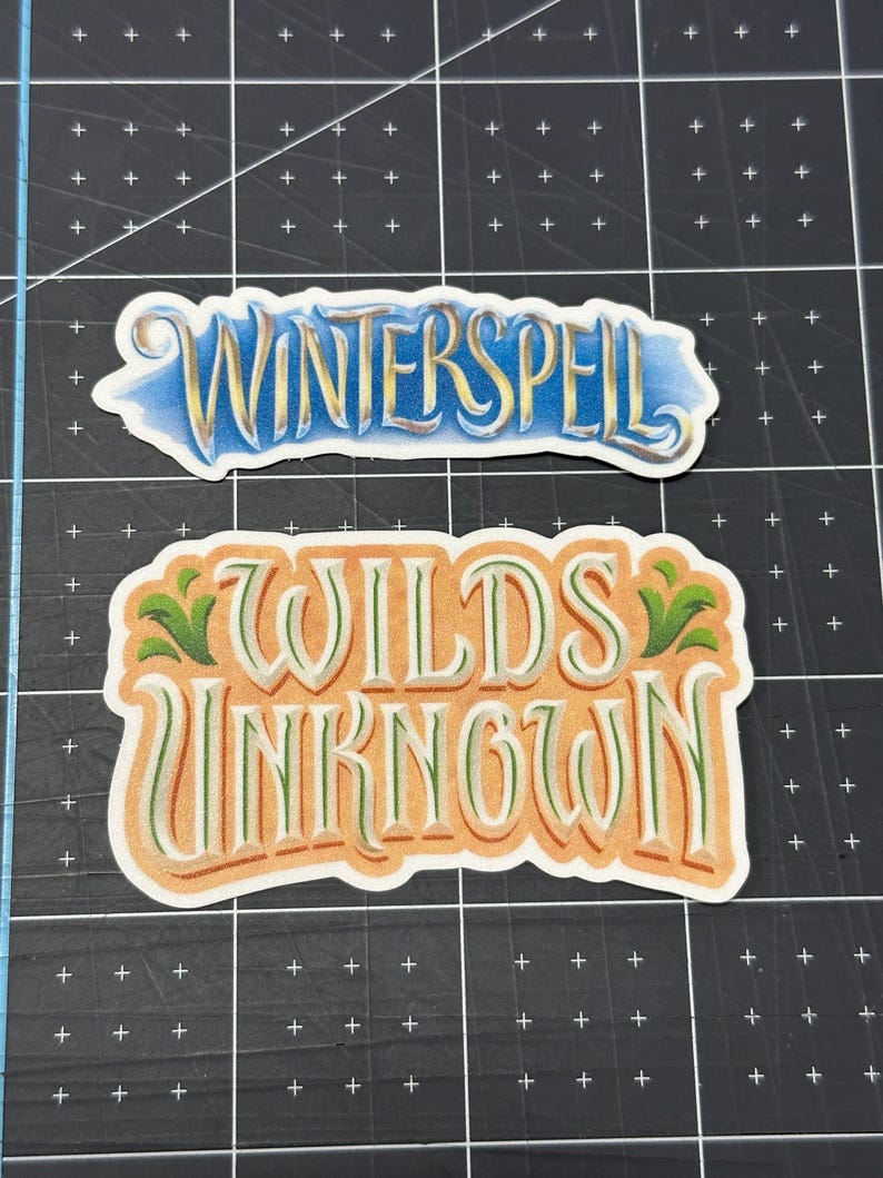 May include: Two decorative stickers with text. The top sticker reads "WINTERSPELL" in gold lettering with a blue and yellow background. The bottom sticker reads "WILDS UNKNOWN" in orange and green lettering.