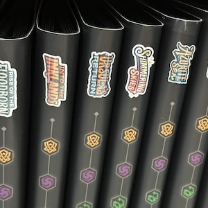 May include: A row of black binders with colorful stickers. The stickers display text such as "Rise of the Floodborn", "Into the Inklands", and "Unbound Return". Each binder has a geometric design in gold and purple.