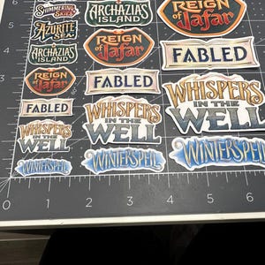 May include: A collection of colorful stickers with fantasy-themed text. The stickers feature phrases like "Whispers in the Well," "Winterspell," and "Reign of Jafar." The stickers are on a black surface with measurement markings.