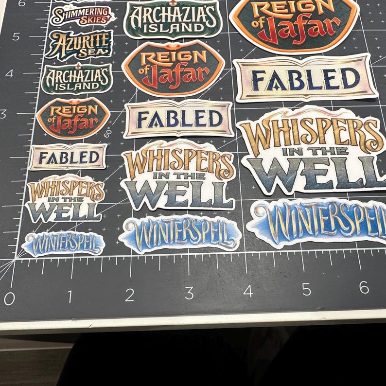 May include: A collection of colorful stickers with fantasy-themed text. The stickers feature phrases like "Whispers in the Well," "Winterspell," and "Reign of Jafar." The stickers are on a black surface with measurement markings.
