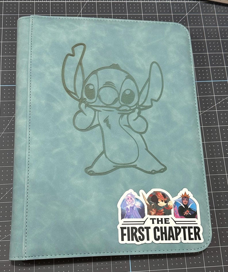 K&ouml;nnte beinhalten: Ein blaugr&uuml;ner Ordner mit einem gen&auml;hten Rand und einem hellblauen, gepr&auml;gten Bild von Stitch aus Lilo & Stitch. Der Ordner hat einen Aufkleber in der unteren rechten Ecke, auf dem "The First Chapter" steht, mit Bildern von drei Disney-Charakteren.