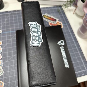 May include: Black card binder with a "RISE OF THE FLOODBORN" sticker. A second black card binder is visible behind the first. A black card holder with the "CARDGUARD" logo is also visible. The binders are on a gray cutting mat.