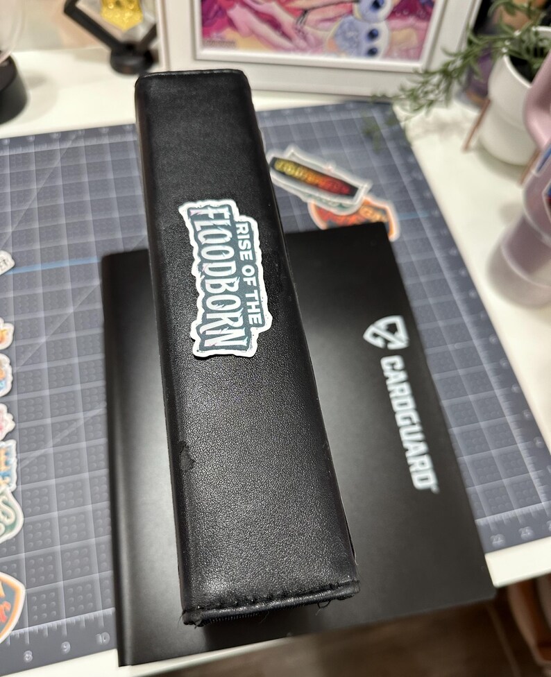 May include: Black card binder with a "RISE OF THE FLOODBORN" sticker. A second black card binder is visible behind the first. A black card holder with the "CARDGUARD" logo is also visible. The binders are on a gray cutting mat.