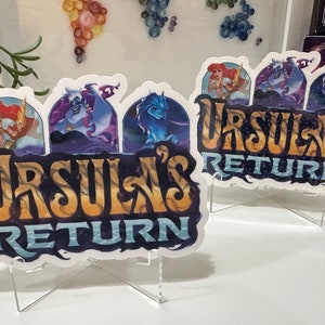 K&ouml;nnte beinhalten: Zwei wei&szlig;e Aufkleber mit einem blauen, violetten und goldenen Design. Die Aufkleber zeigen den Text "Ursula's Return" und Bilder von Ursula, einer Meerjungfrau und einer Meereskreatur. Die Aufkleber befinden sich auf klaren Acrylst&auml;ndern.